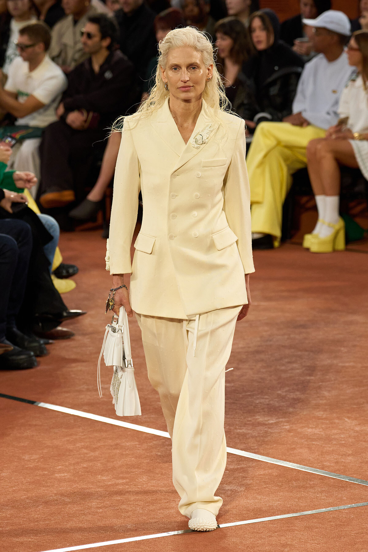 Model walking the runway in a cream double-breasted suit with a matching bag as the audience watches.