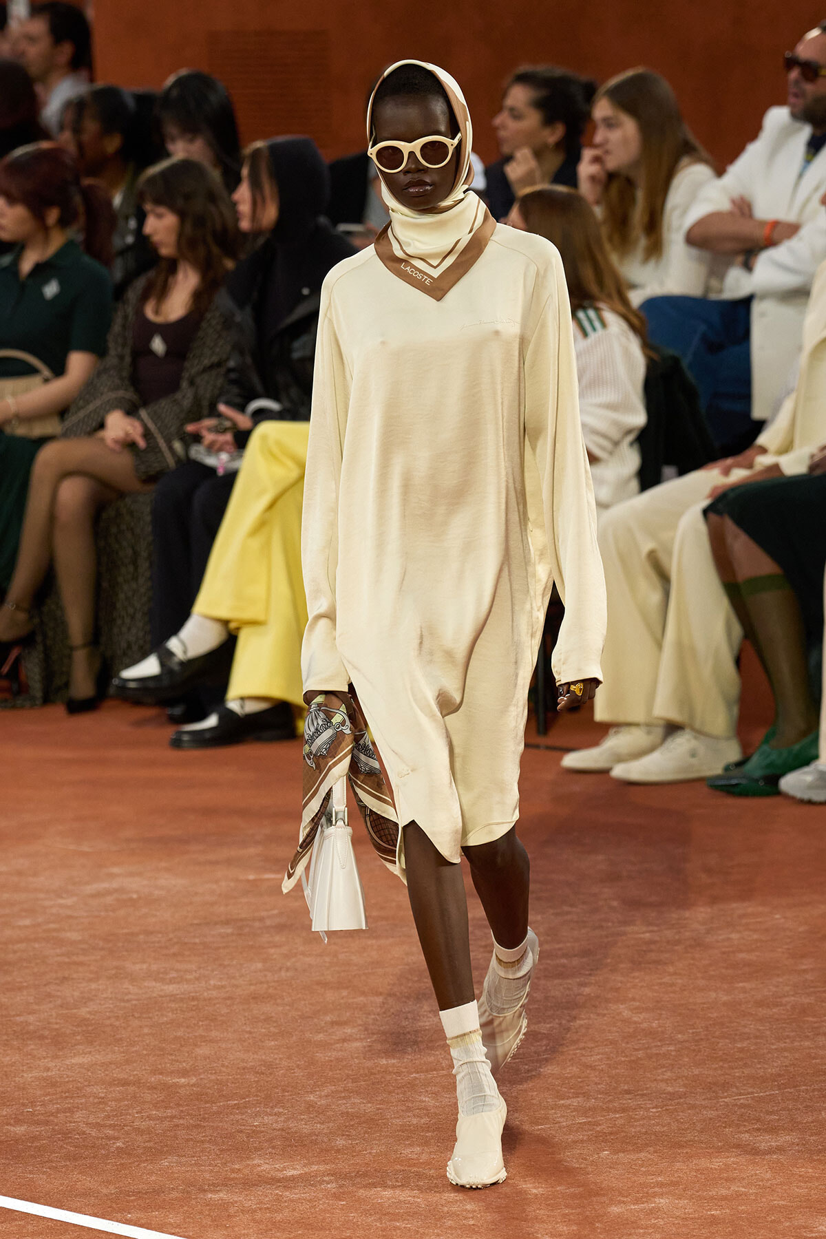 Model on a runway wearing a loose cream dress with a matching headscarf and oversized white sunglasses, carrying a small white bag and a patterned scarf draped at the side.