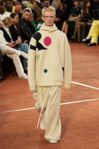 Model walks the runway in an oversized cream sweater with bold geometric shapes and matching wide-leg pants, audiences seated in the background.
