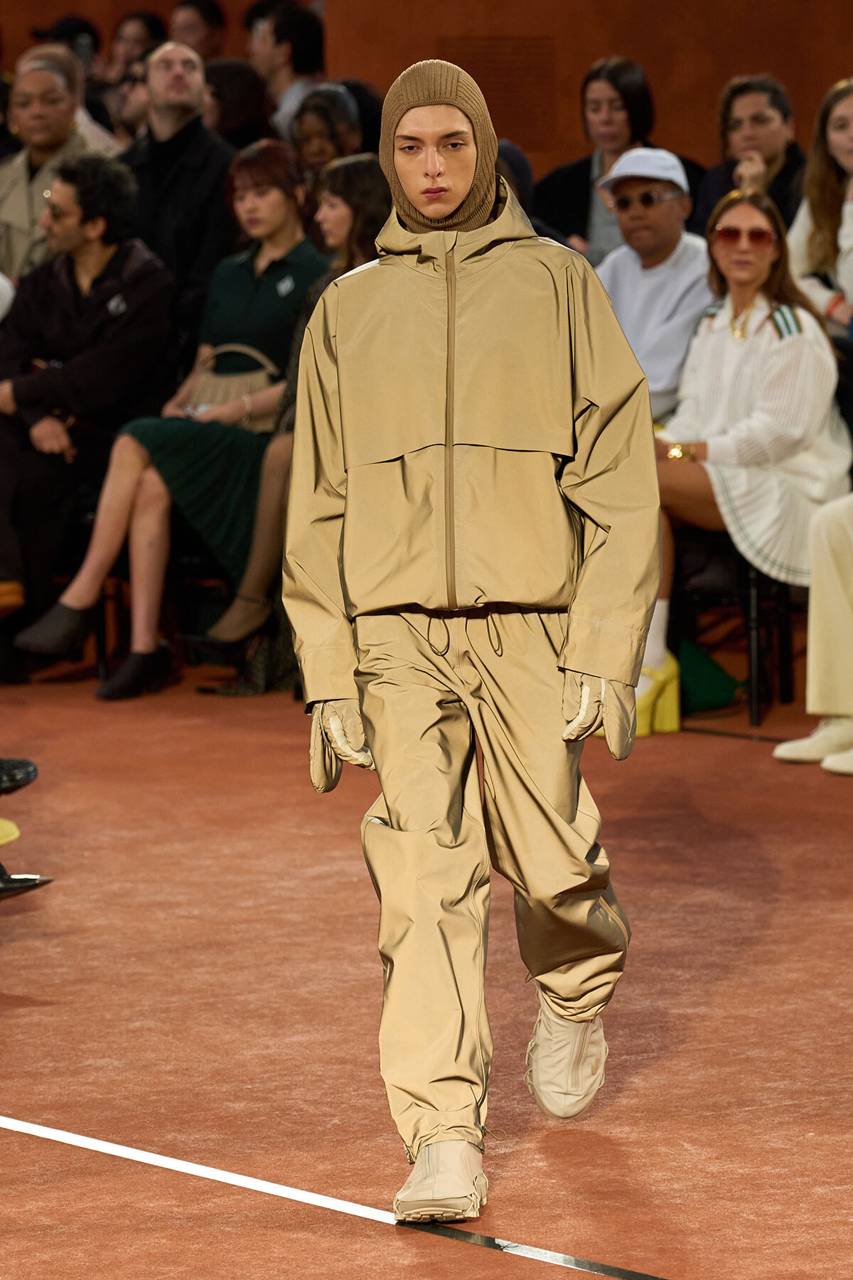 Model walks the runway in a beige hooded windbreaker and matching pants, wearing gloves, with an audience in the background.