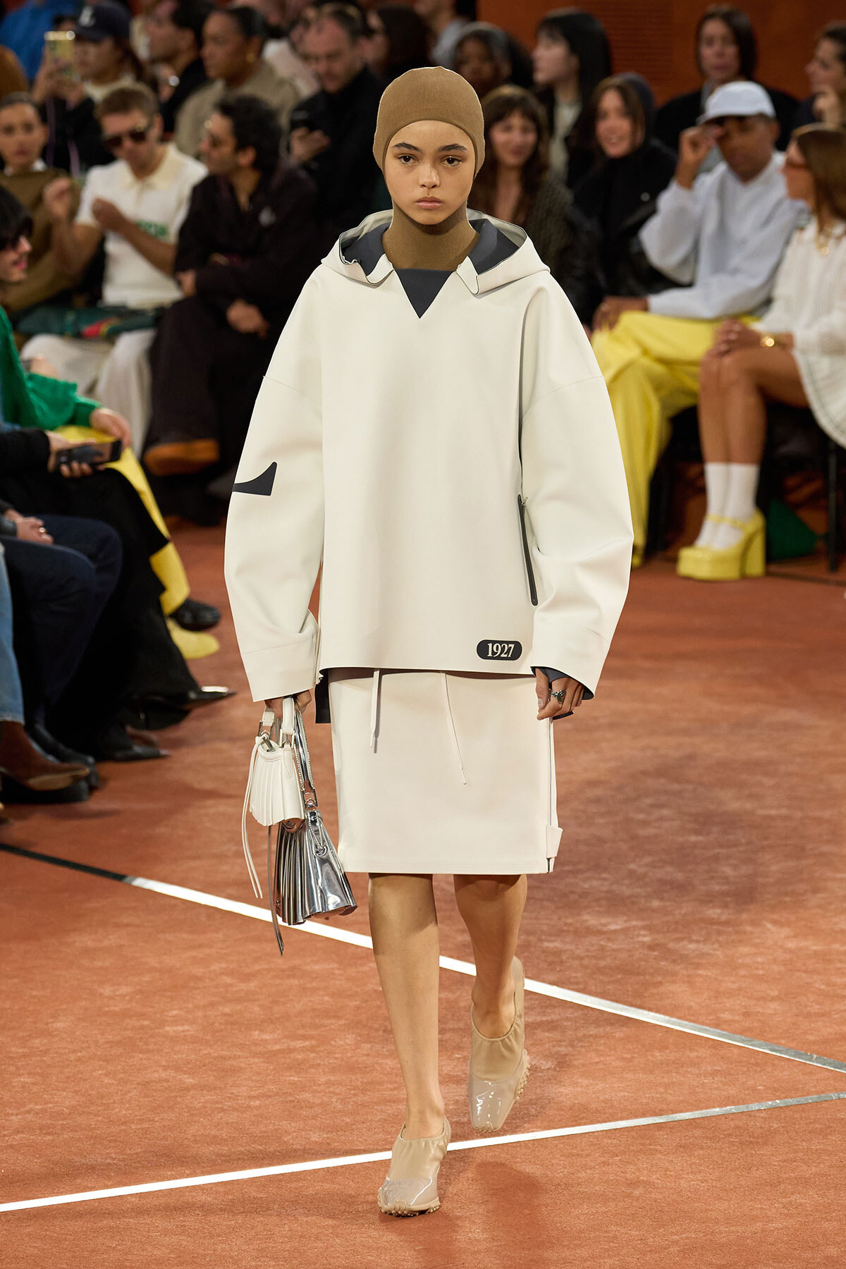 Model walks the runway in a minimalist white oversized coat with a beige headscarf, carrying metallic handbags.