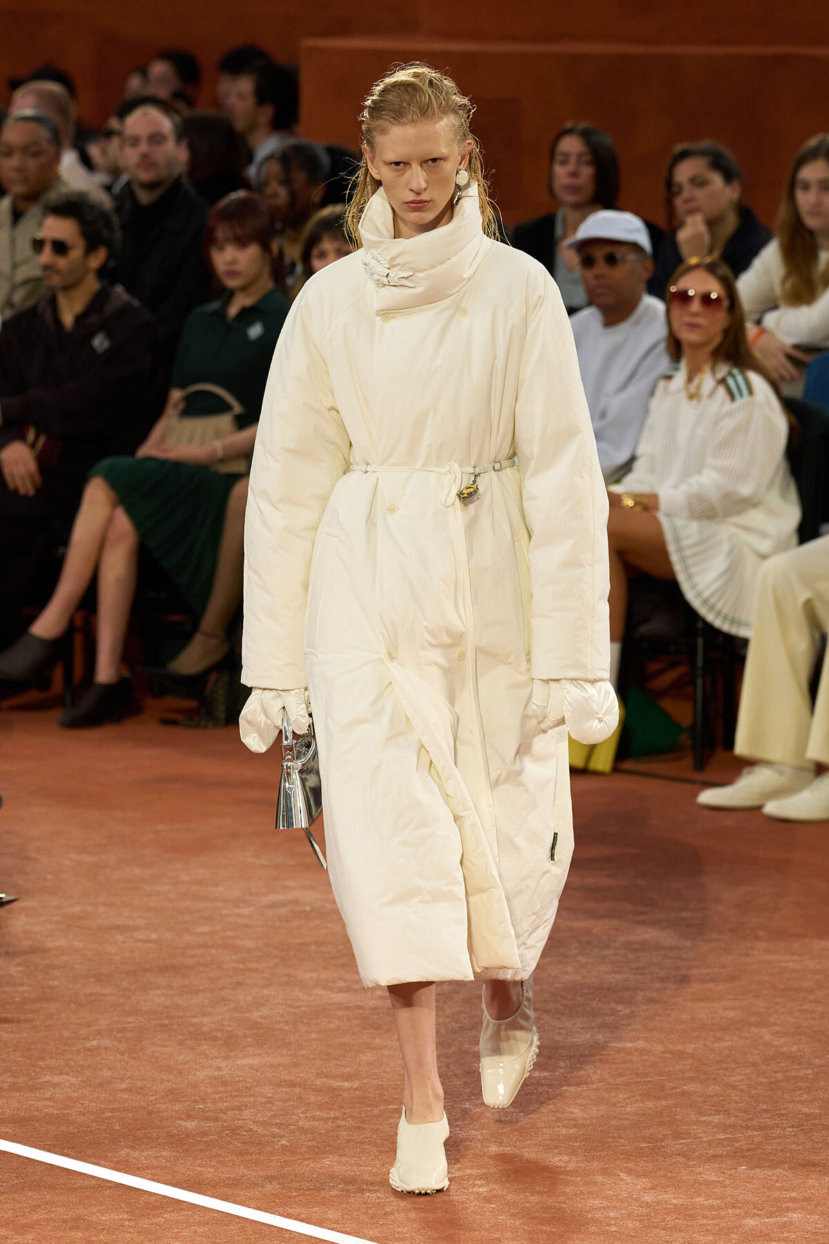 Model walks the runway in a long white quilted coat with high collar and belt, audience blurred in the background.