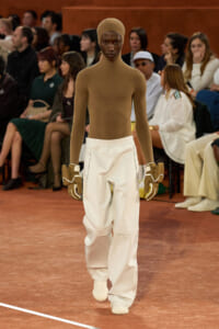 Model walking a runway in a taupe full-body knit suit with matching cap, white oversized trousers, and gloves with white trim; audience in the background.