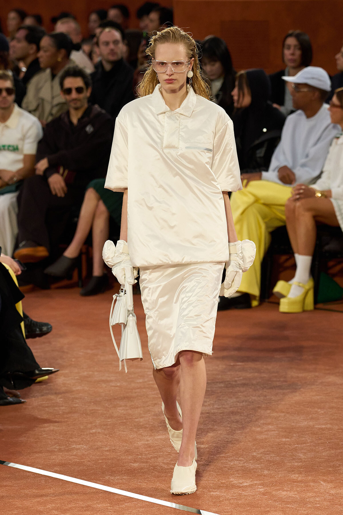 Model walks a runway in an ivory satin shirt-dress with short sleeves, matching gloves, and a white handbag.
