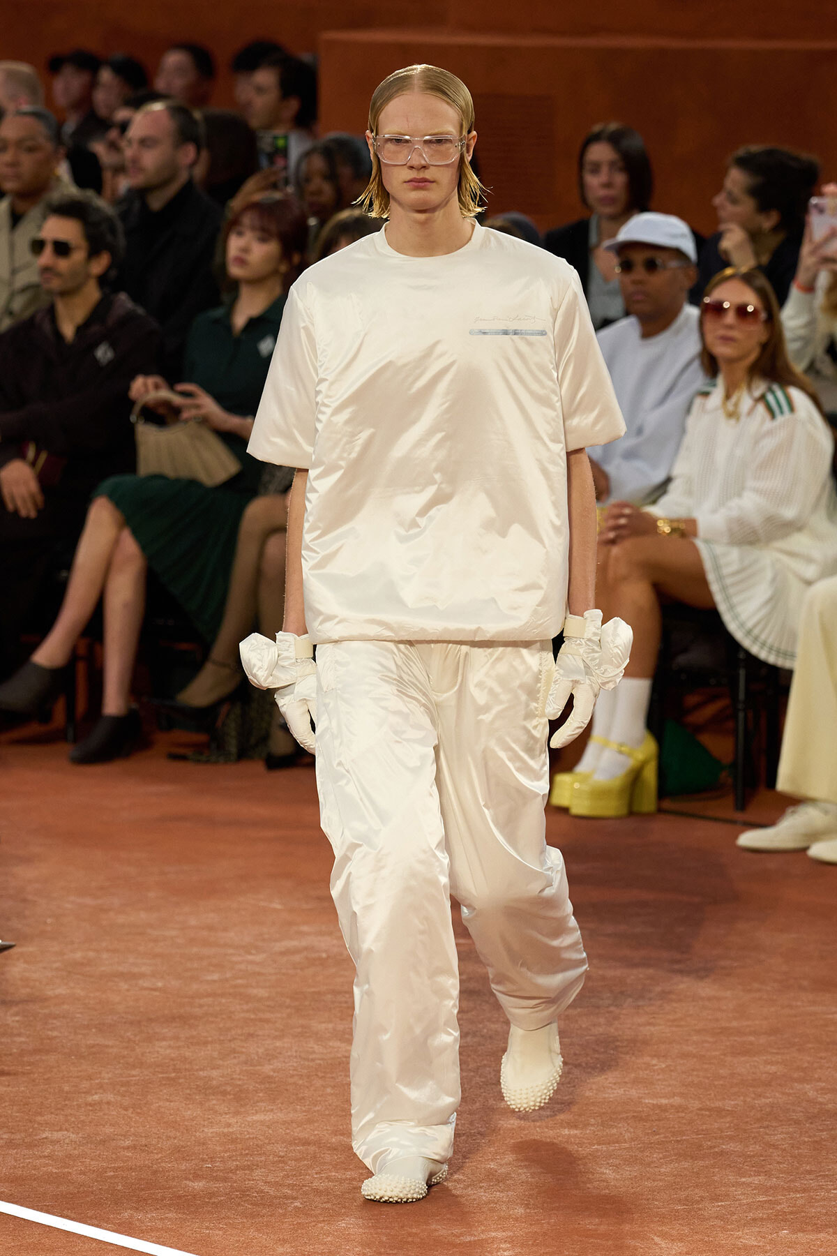 Model walks a fashion show in an all-white satin outfit with gloves and protective eyewear, audience watching.