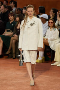 Model on a runway wearing a white double-breasted coat and matching shorts, holding a metallic handbag, with seated audience in background.