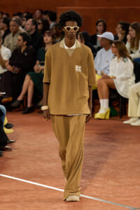 Male model in a camel-tan oversized tracksuit with matching wide trousers and white sunglasses walking a fashion runway, audience in background.