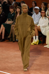 Model walks the runway in a tan utility jumpsuit with a matching headscarf; audience sits in the background.