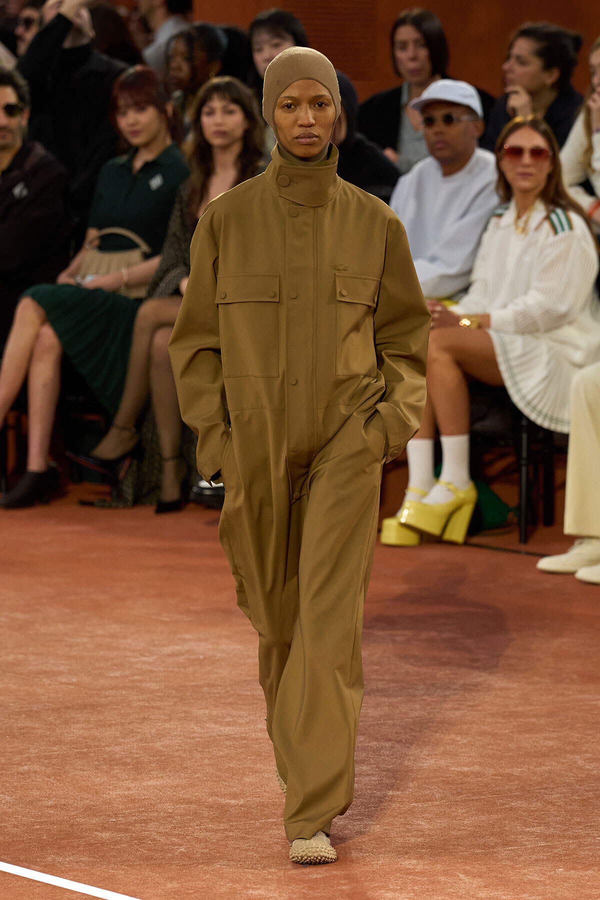 Model walks the runway in a tan utility jumpsuit with a matching headscarf; audience sits in the background.
