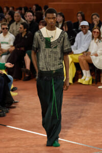 Model walks a fashion runway wearing a sheer embroidered top over a green shirt and loose dark green trousers, with layered necklaces; audience seated along the sides.