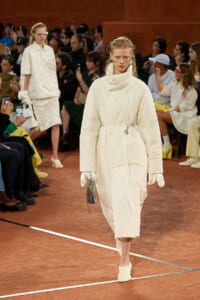 Model walks a cream oversized quilted coat with belt on a fashion runway, audience watching