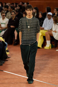 Model walks the runway in a black short-sleeve top with metallic square embellishments and loose dark green pants, accessorized with layered necklaces.