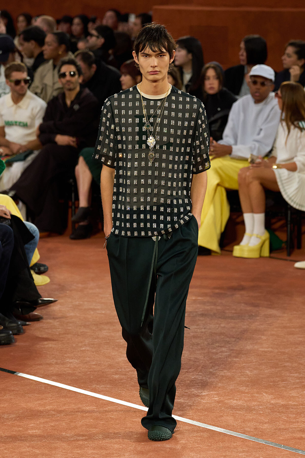 Model walks the runway in a black short-sleeve top with metallic square embellishments and loose dark green pants, accessorized with layered necklaces.