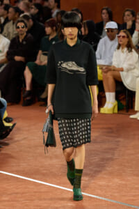 Model walks a runway in a black oversized sweatshirt with a silver sparkle sock/shoe graphic, paired with patterned black shorts, green socks, and green shoes, carrying a dark green handbag; seated audience in background.
