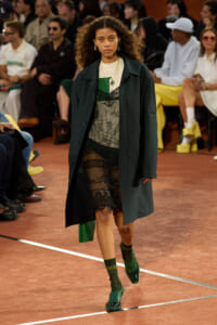 Model walks the runway in a dark green coat over a sheer lace dress, with layered necklaces and green shoes, audience in the background.