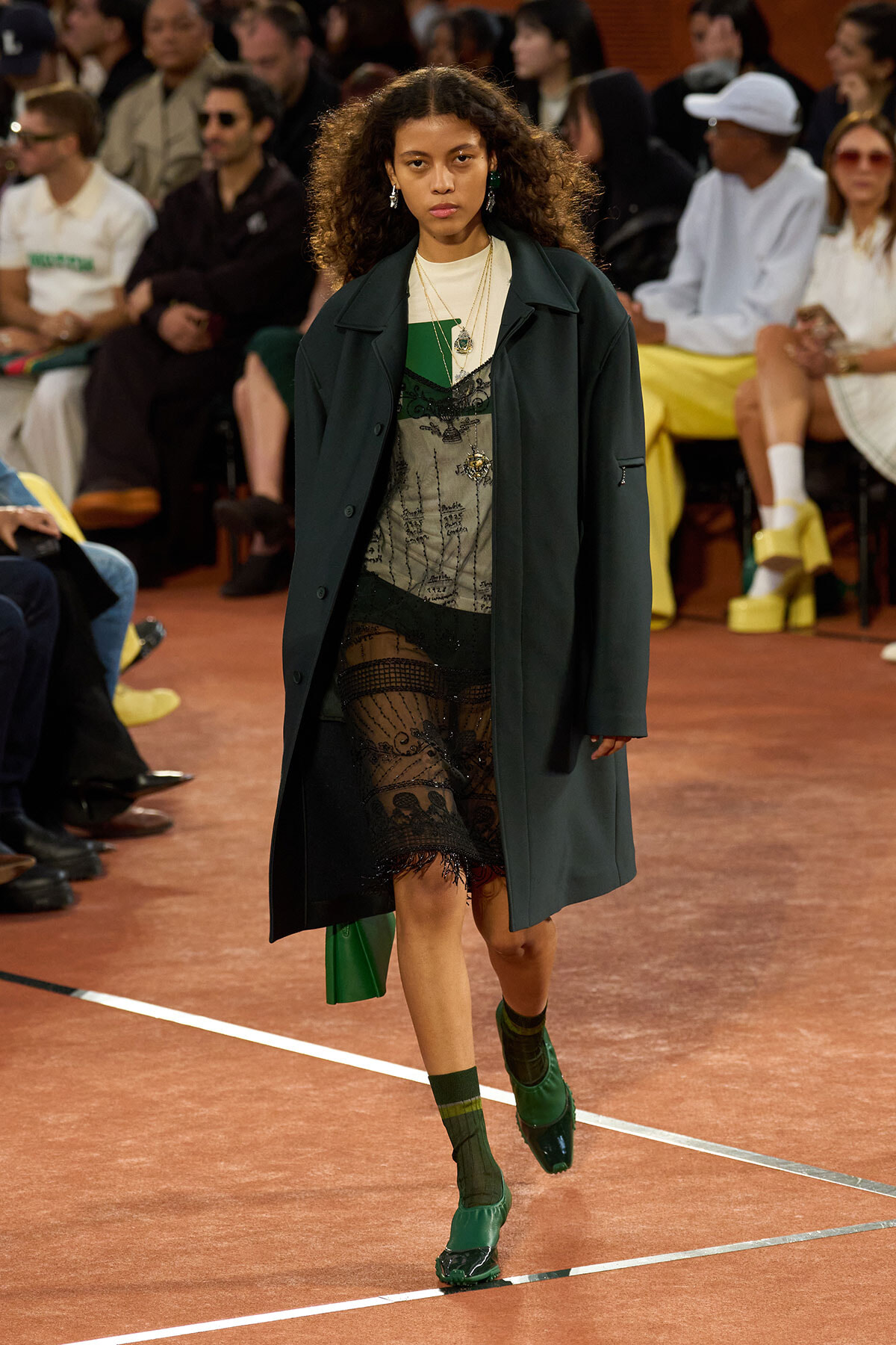 Model walks the runway in a dark green coat over a sheer lace dress, with layered necklaces and green shoes, audience in the background.