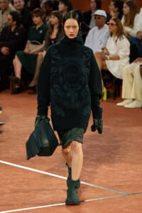 Model walking a runway in a dark green oversized turtleneck sweater with a Lacoste crest, paired with green gloves, a green geometric bag, and matching boots while watching an audience.