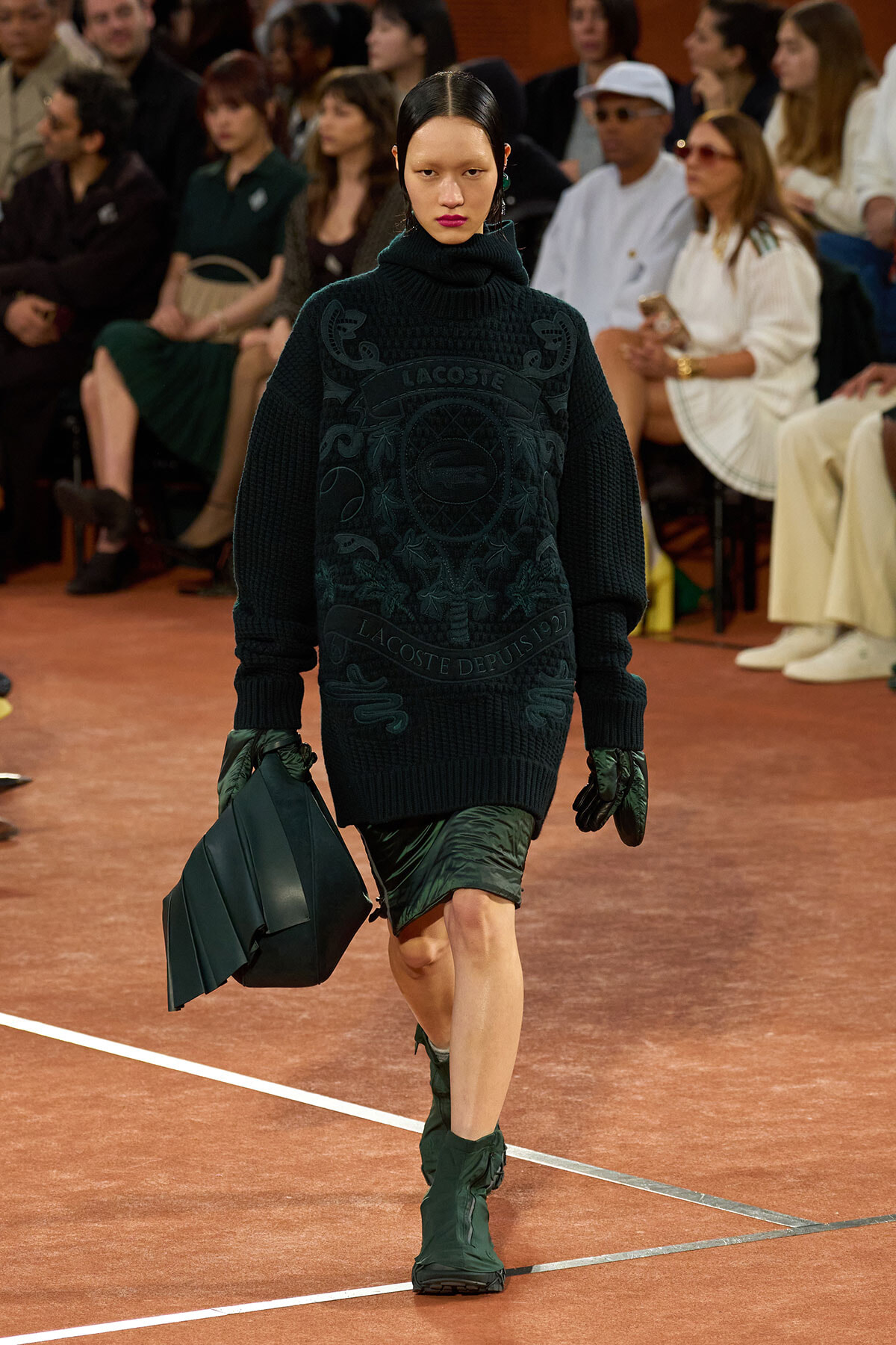 Model walking a runway in a dark green oversized turtleneck sweater with a Lacoste crest, paired with green gloves, a green geometric bag, and matching boots while watching an audience.