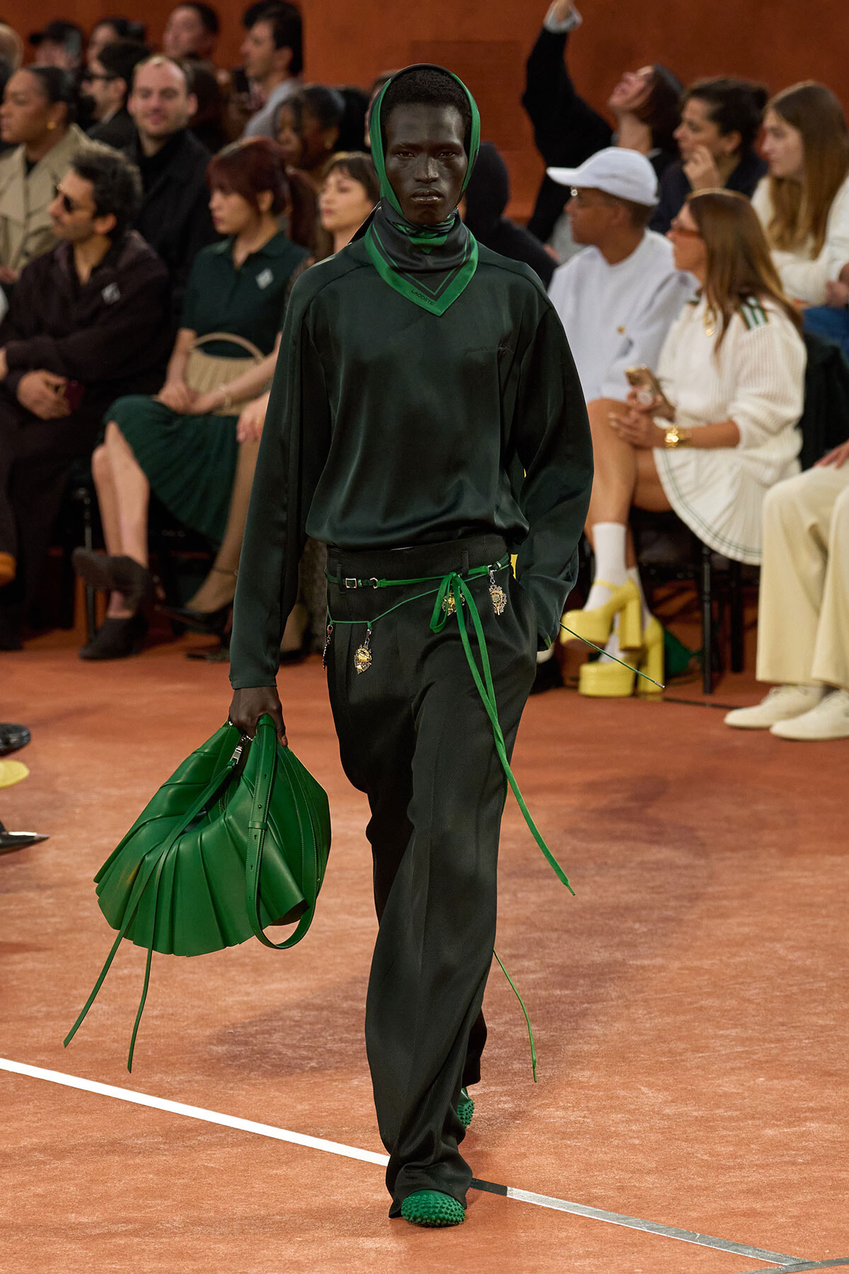 Model walks a runway in an all-green outfit with a green scarf and oversized green handbag in hand