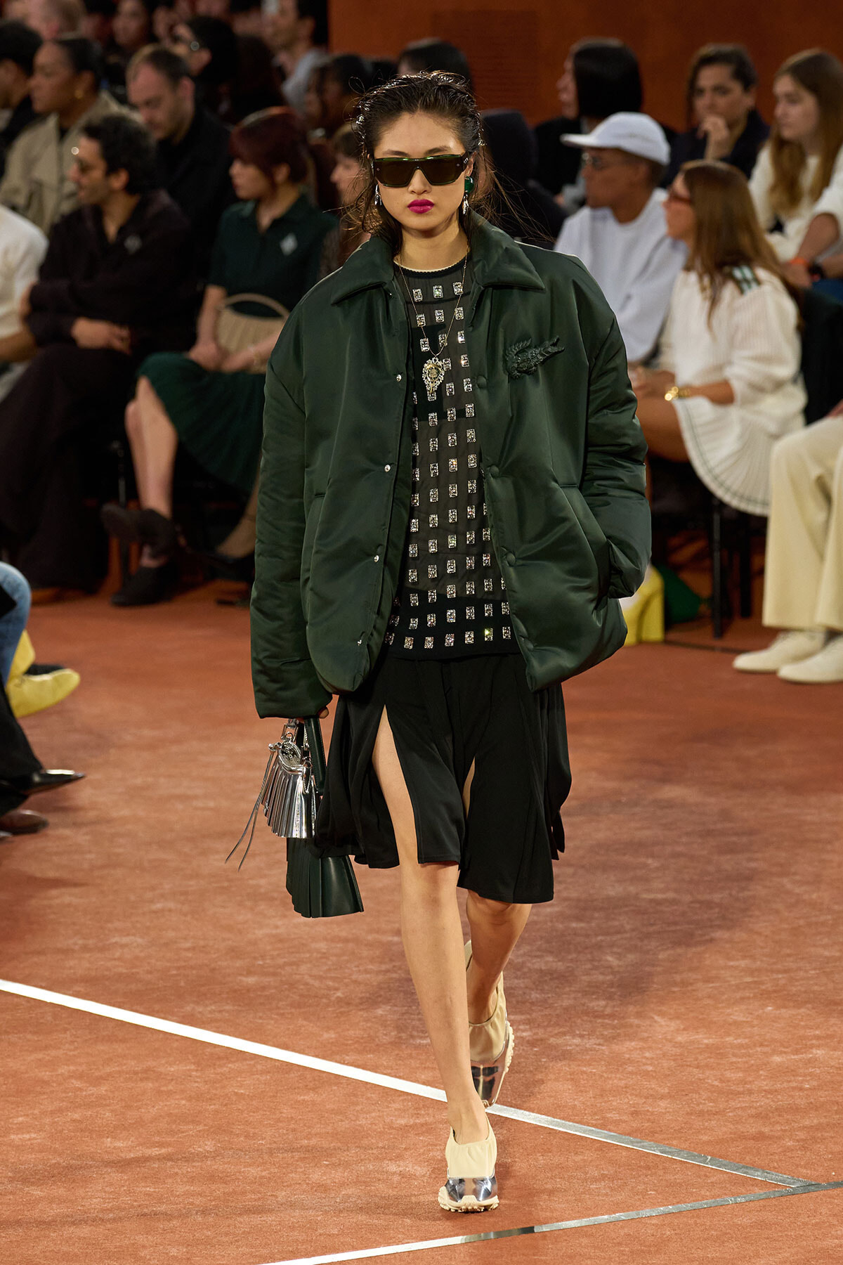 Model walks the runway in a dark green oversized puffer jacket over a black square-patterned dress with high side slits; large sunglasses and metallic shoes.