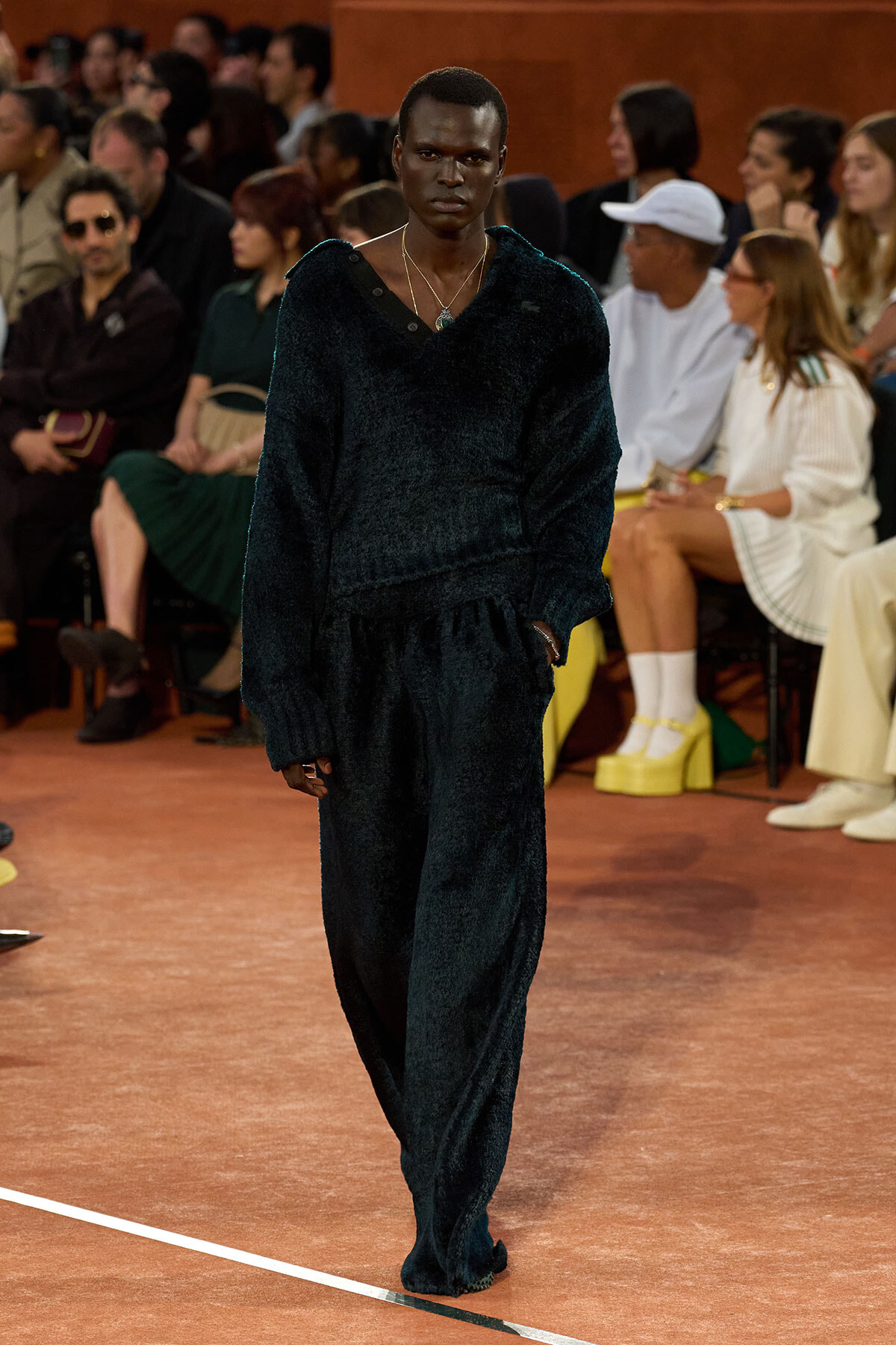 Model walks the runway in a matching dark velvet loungewear set with layered necklaces; audience seated behind.