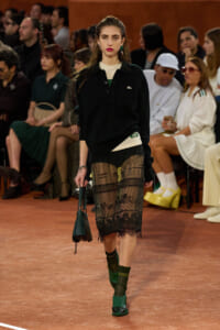 Model walking a runway in a black polo sweater layered over a beige top, and a sheer black lace skirt with writing, paired with green platform shoes and a small green handbag, while an audience watches.