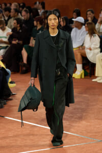 Model walking a runway in a dark green coat and matching pants, carrying a structured green bag; audience visible in background.