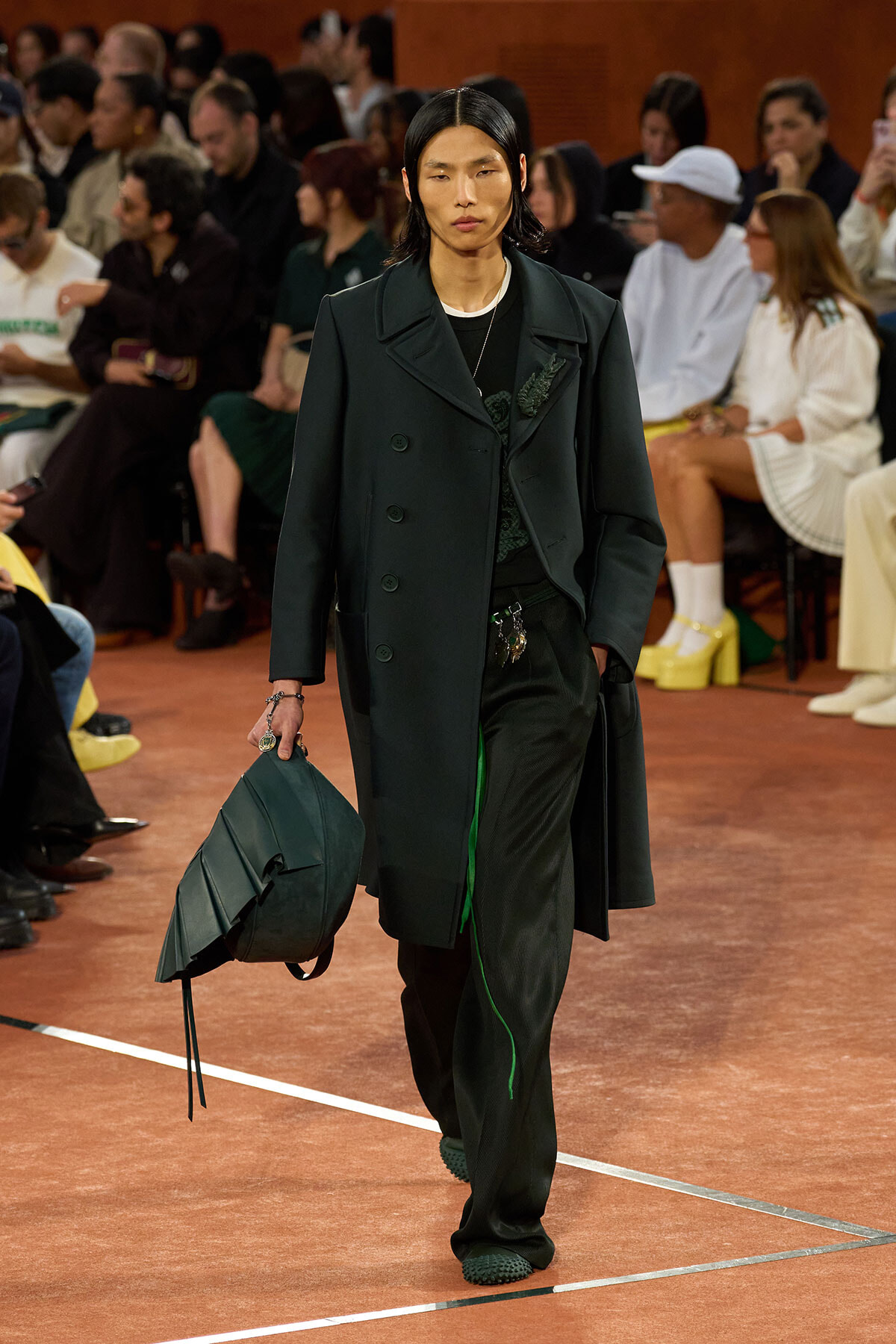 Model walking a runway in a dark green coat and matching pants, carrying a structured green bag; audience visible in background.