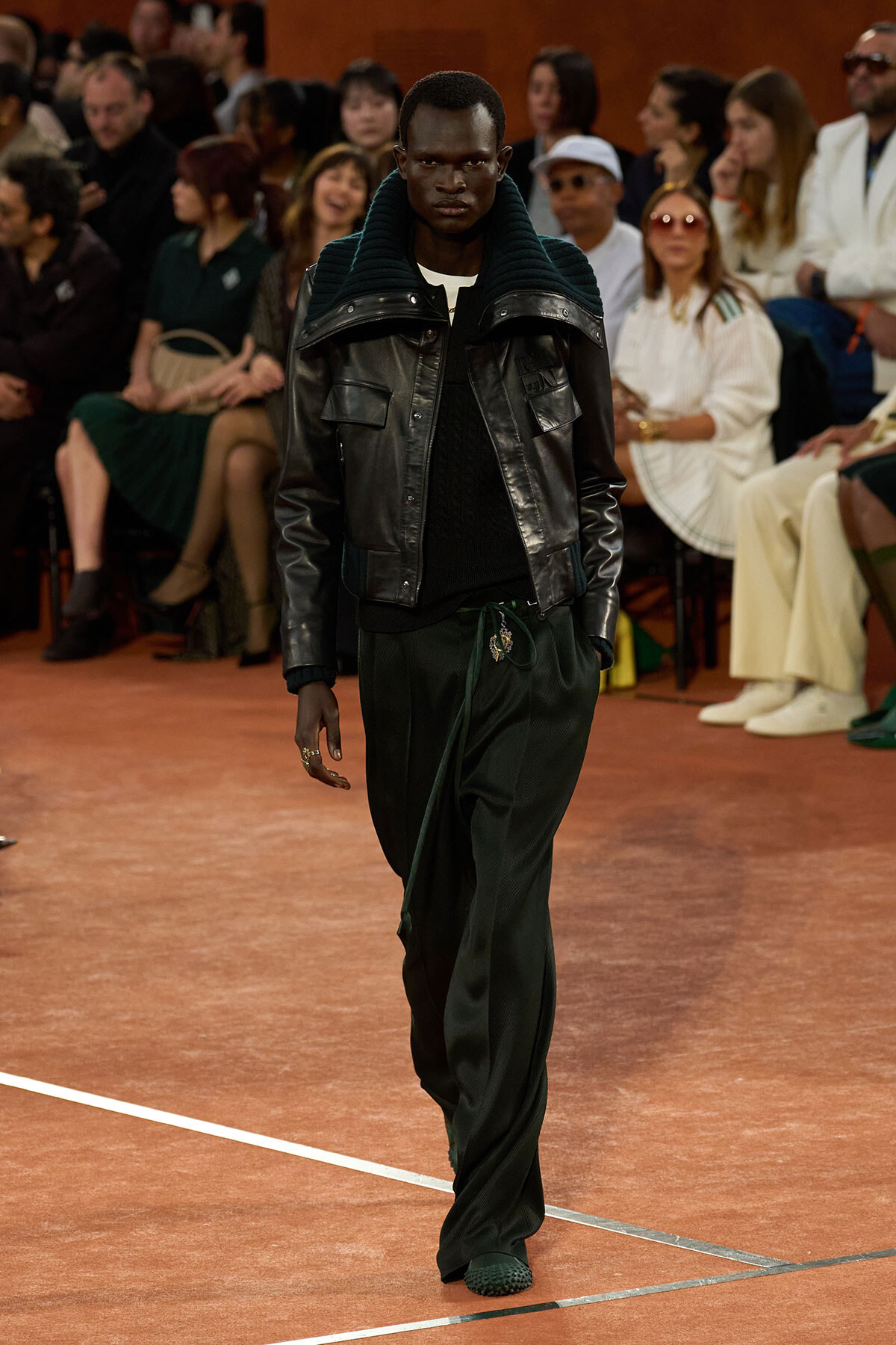 Male model walking down a fashion runway in a black leather jacket with a high dark teal collar, black sweater, and loose dark green pants; audience seated in the background.
