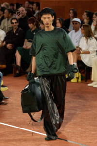 Male model on a fashion runway in a dark green oversized top and black leather pants, carrying a large dark green duffel bag.