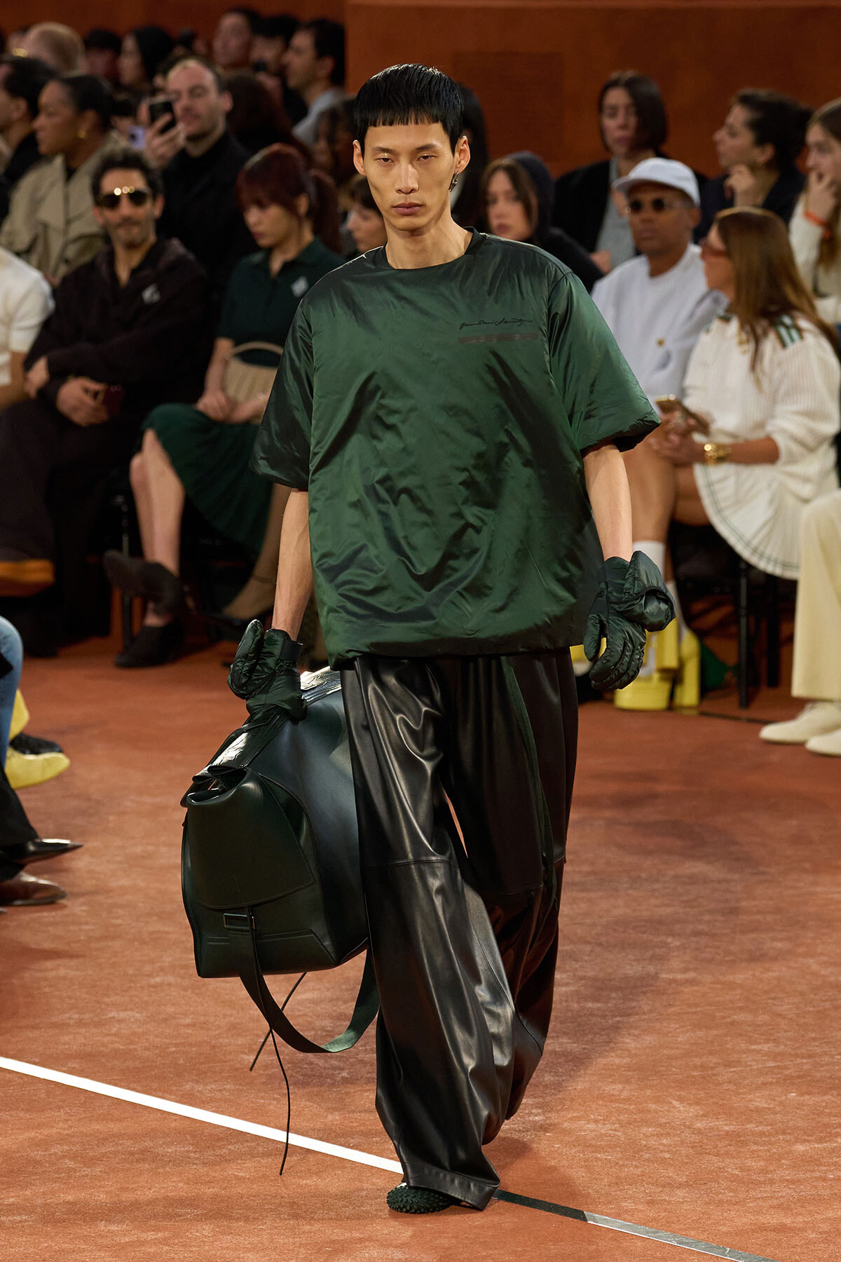 Male model on a fashion runway in a dark green oversized top and black leather pants, carrying a large dark green duffel bag.