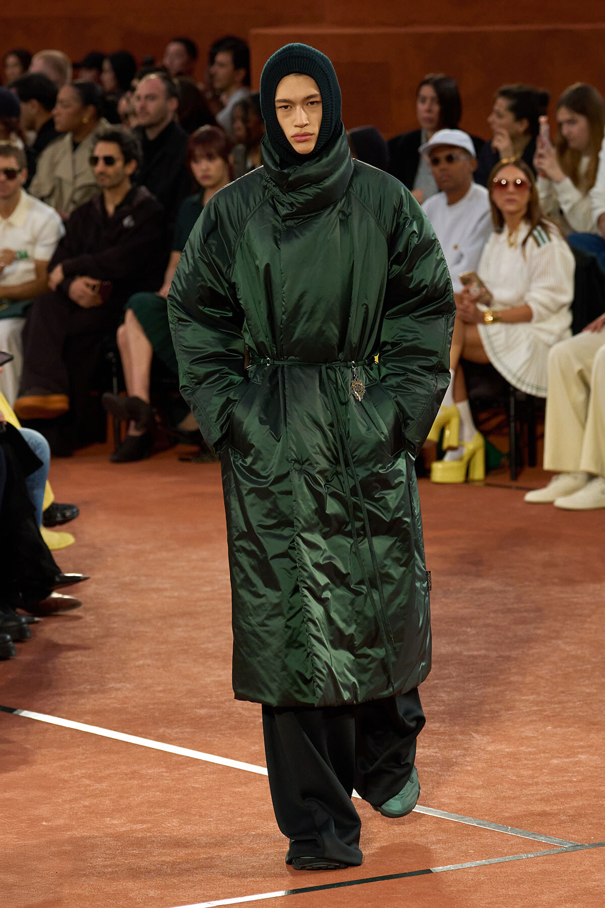 Model on a runway wearing a dark green, shiny belted puffer coat with a hood and a knit cap, audience in the background.