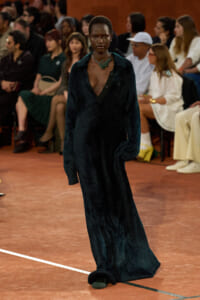 Model walks the runway in a long dark plush coat with a deep V neckline and green statement necklace, with seated audience in the background to the side.