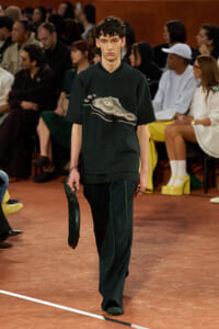 Male model walks a fashion runway in a dark green top with a jeweled crocodile graphic, matching wide-leg pants, and a dark green clutch; audience sits along the catwalk in the background.