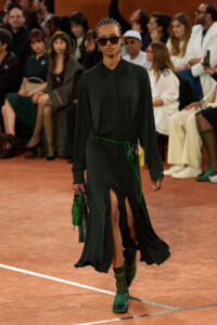 Model walks a runway in a dark green long-sleeve dress with a black lace collar, green belt, and green accessories, carrying a green handbag; audience watching.