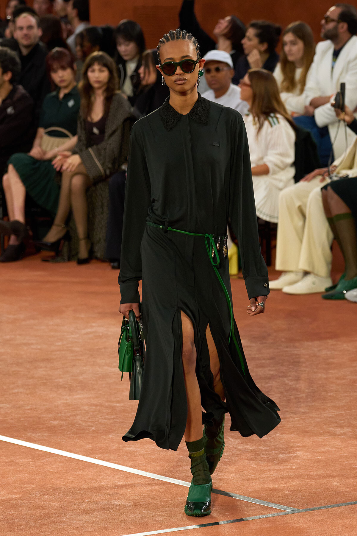 Model walks a runway in a dark green long-sleeve dress with a black lace collar, green belt, and green accessories, carrying a green handbag; audience watching.