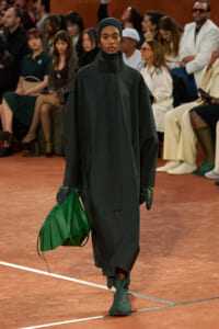 Model on a runway wearing a dark green cape and matching headscarf, with green gloves and boots, carrying a bright green handbag; audience in background.