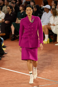 Model walking a fashion runway in a bright magenta double-breasted blazer with matching knee-length skirt, jeweled brooch at the lapel, clear protective glasses, and white socks with white shoes; audience seated in the background.