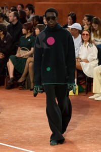 Model walks a runway in an oversized dark sweater with bright pink and green circular patches, paired with gloves and sunglasses, audience in the background.
