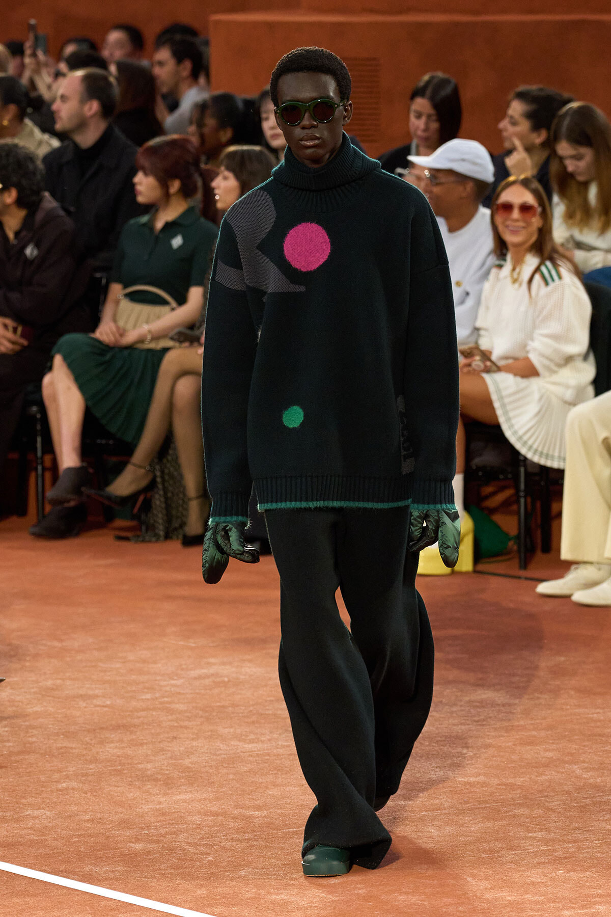 Model walks a runway in an oversized dark sweater with bright pink and green circular patches, paired with gloves and sunglasses, audience in the background.