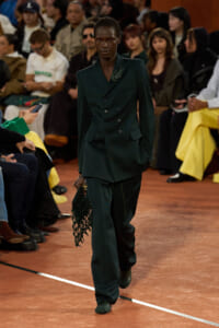 Model walking the runway in a dark emerald green double-breasted suit with a beaded bag, audience in the background.
