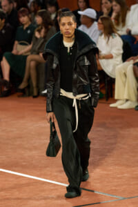 Model walking a fashion runway in a black leather jacket with teal knit collar, black sweater, and dark green wide-leg pants; carrying a small green handbag.