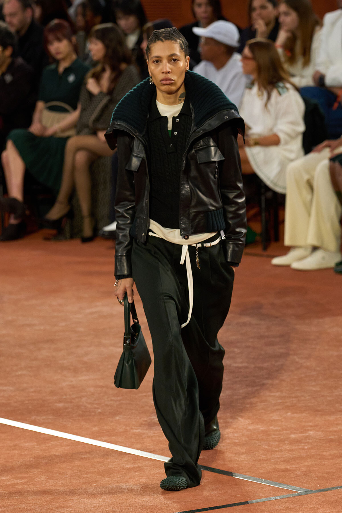 Model walking a fashion runway in a black leather jacket with teal knit collar, black sweater, and dark green wide-leg pants; carrying a small green handbag.