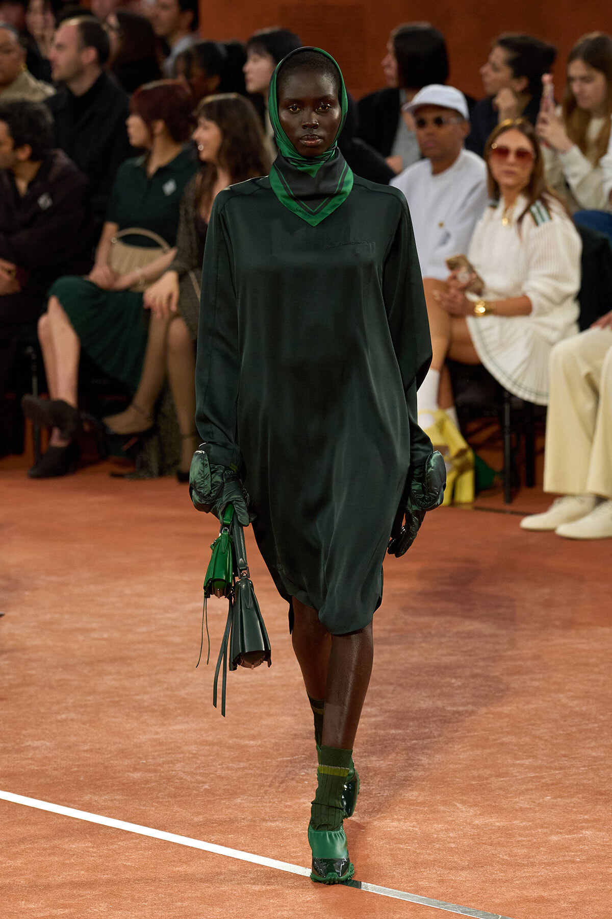 Model in a dark green satin dress and matching hijab walks the runway, wearing oversized gloves and carrying green handbags as spectators watch from the sides.