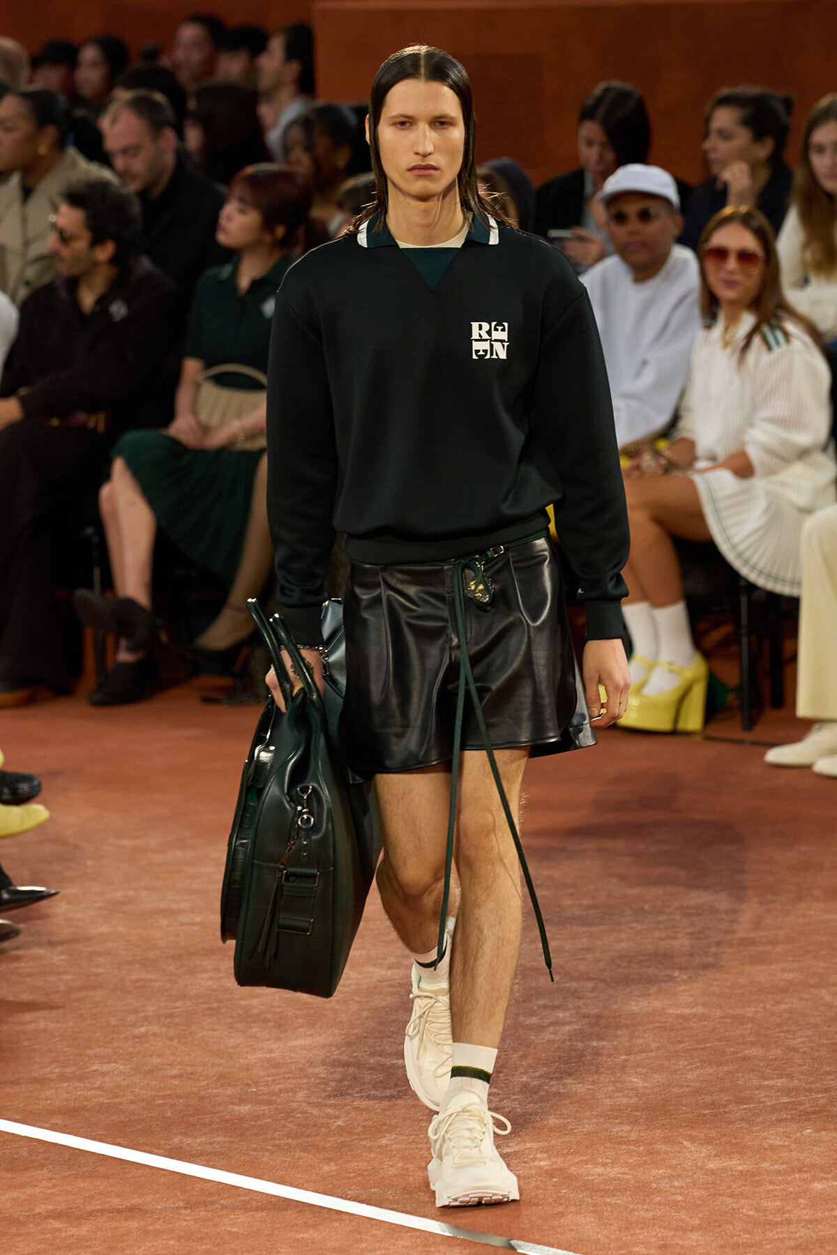 A male model walks the runway wearing a black sweatshirt and black leather mini skirt, carrying a large dark green bag.