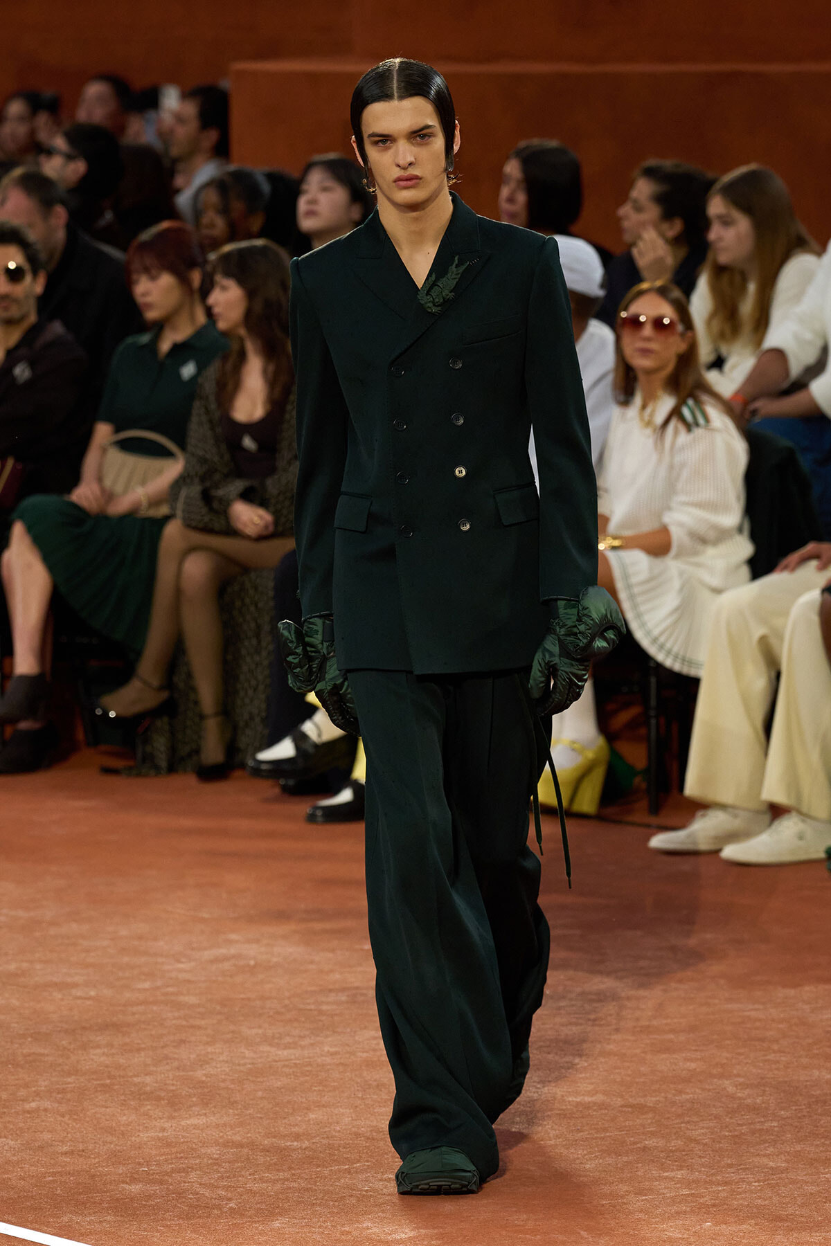 Model walking the runway in a dark green double-breasted suit with quilted gloves, audience in the background.