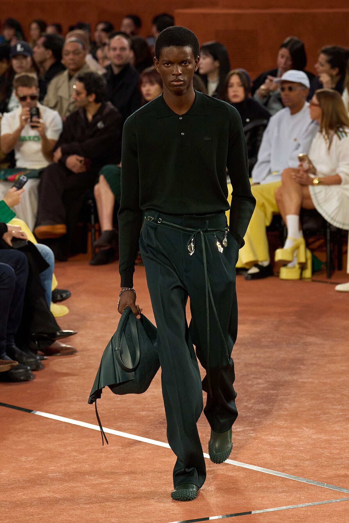 Male model walks a runway in a dark green polo and matching wide-leg pants, carrying a large green bag, during a fashion show.