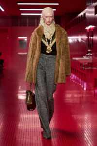 Model walking a fashion show in a brown fur coat, beige headscarf, and layered jewelry in a red-lit hallway.