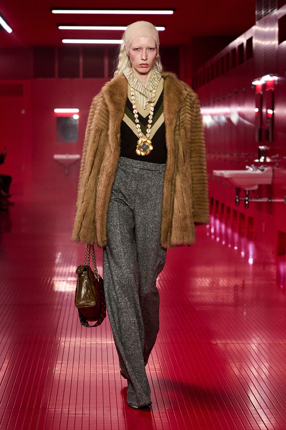 Model walking a fashion show in a brown fur coat, beige headscarf, and layered jewelry in a red-lit hallway.
