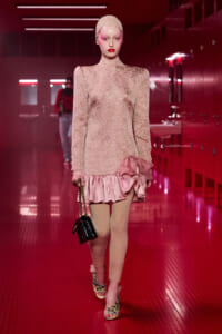 Runway model in a pale pink lace mini dress with a satin ruffle, head wrap, and beige tights, carrying a small black handbag in a pink-lit hallway.